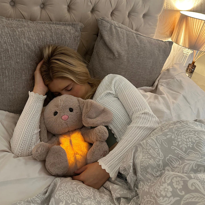 Slapende vrouw in wit gebreid shirt die een pluchen konijn met gloeiende buik omarmt, tussen grijze kussens in bed. 16