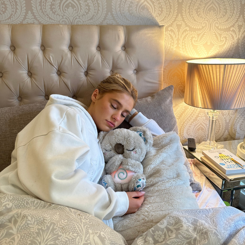 Slapende jongere in witte hoodie die in bed een grijze pluche koala met roze gloeiende spiraal op de buik vasthoudt. 14