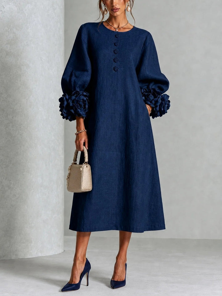 Model draagt de jurk in marineblauw, vooraanzicht, met bloemenknopen, pofmouwen met grote stoffen rozen en een beige handtas. 1