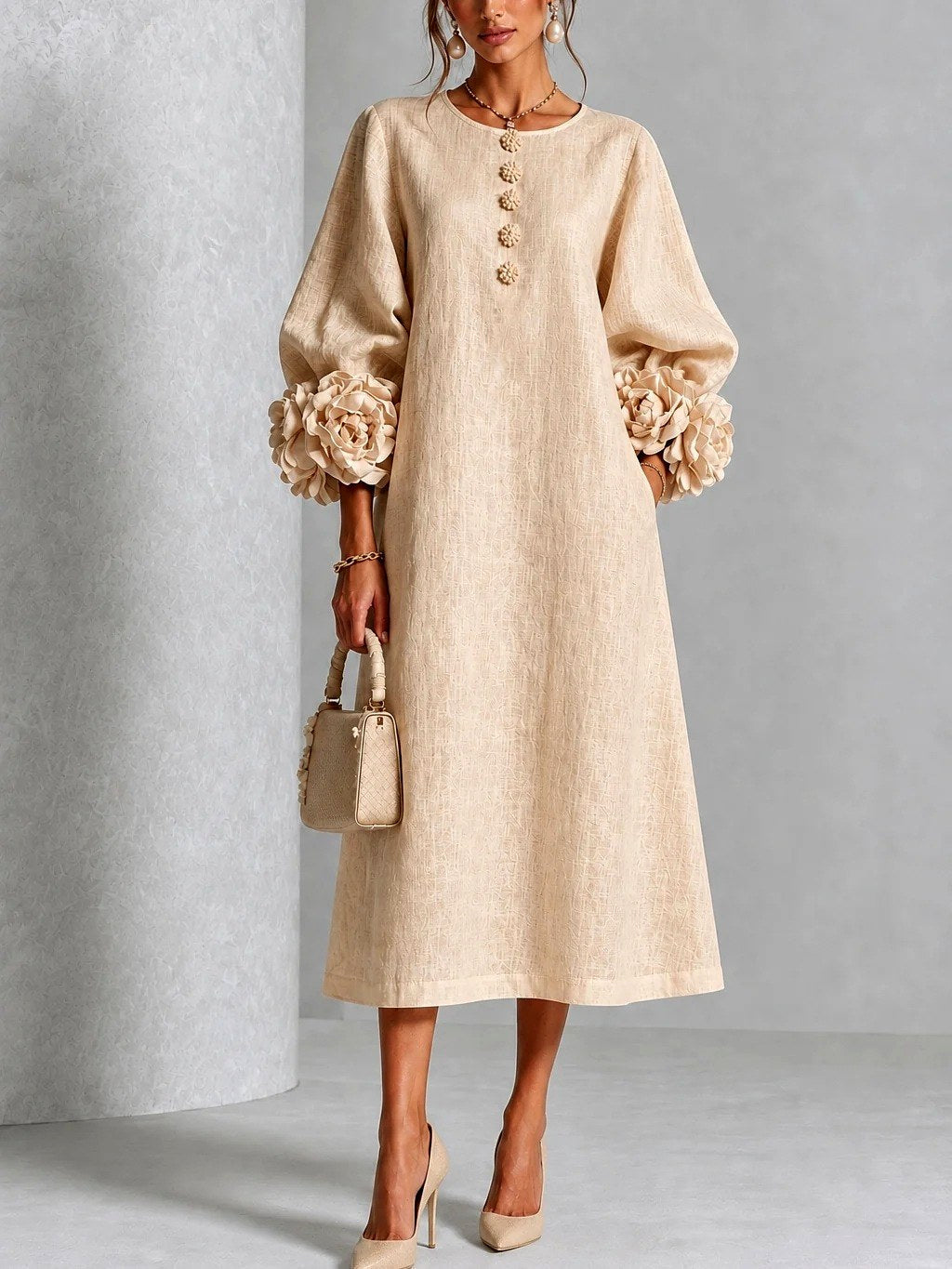 Model draagt de jurk in zandbeige, vooraanzicht, met bloemvormige knopen, wijde A-lijn en rozenmanchetten aan de ruime mouwen. 4