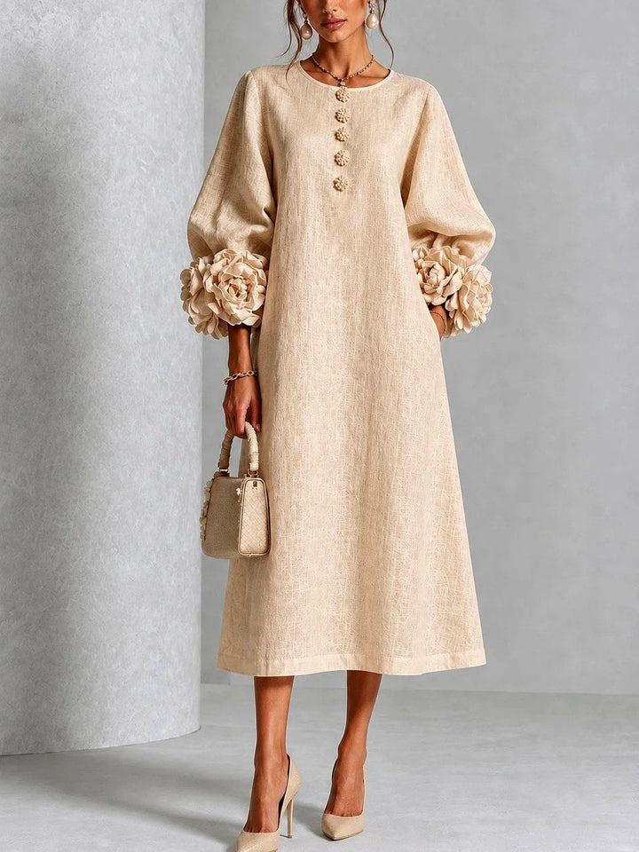 Model draagt de jurk in zandbeige, vooraanzicht, met bloemvormige knopen, wijde A-lijn en rozenmanchetten aan de ruime mouwen. 4