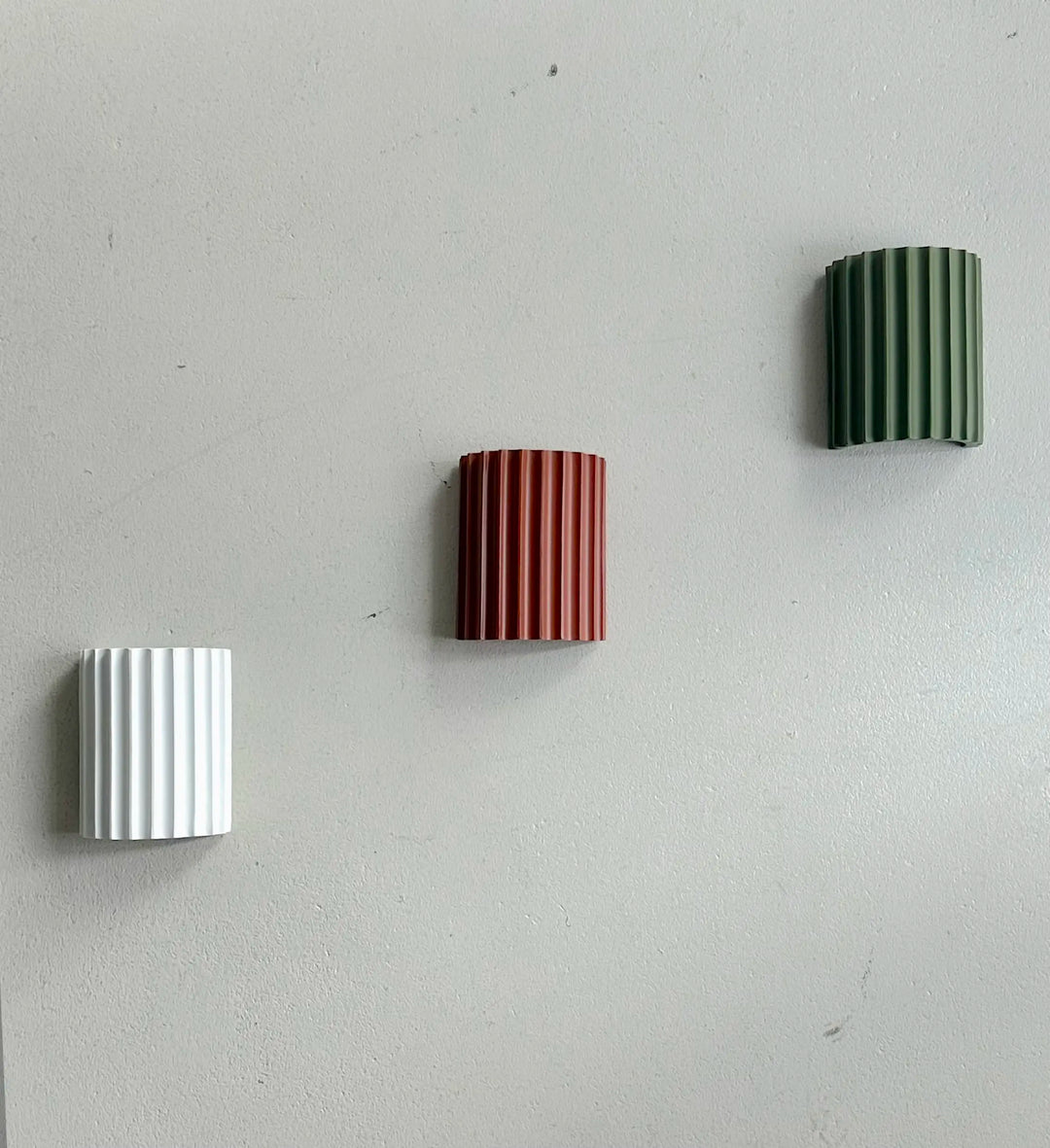 Drie geribbelde wandlampen in wit, terracotta en groen schuin onder elkaar op een grijze muur gemonteerd 12