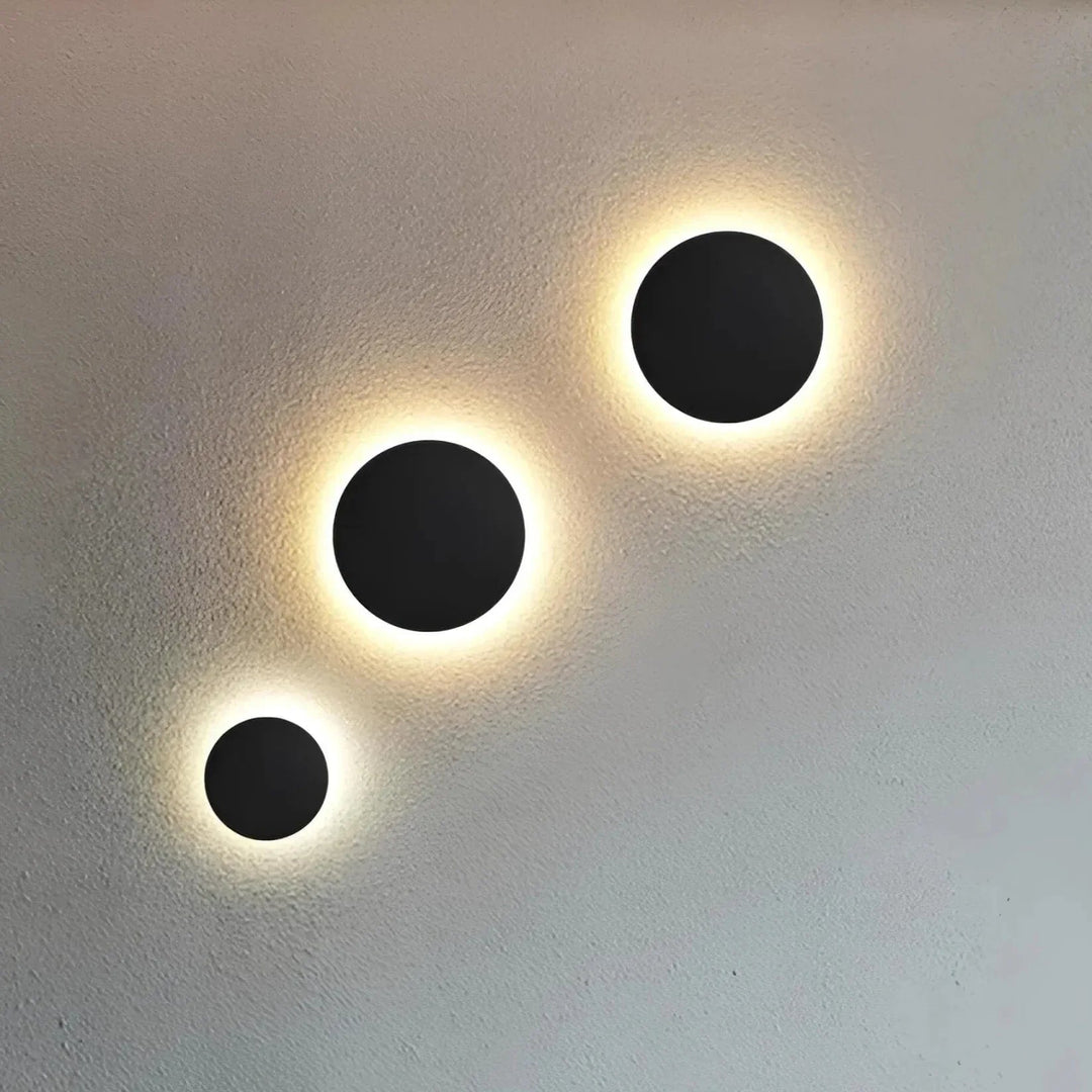 Drie zwarte ronde wandlampen in verschillende maten op een rij, met warm licht dat achter de schijf op de muur straalt. 2