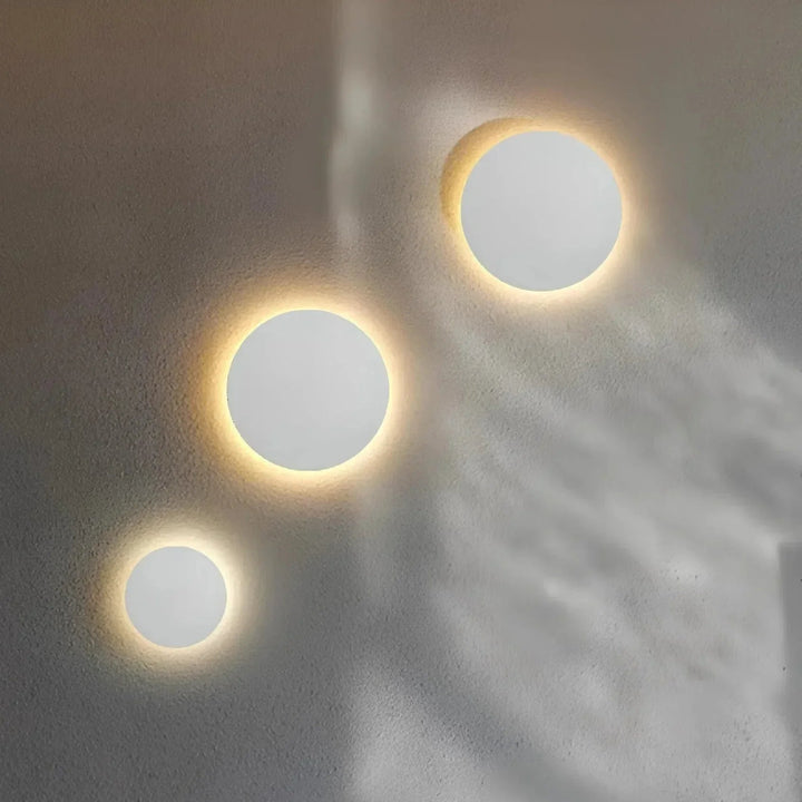 Drie witte ronde wandlampen in verschillende maten op een muur, met warm licht rond de randen. 4