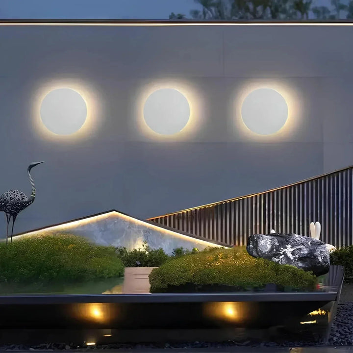 Drie witte ronde wandlampen op rij aan een buitenmuur boven een moderne tuin met mos, rotsen en verlichte randen. 5