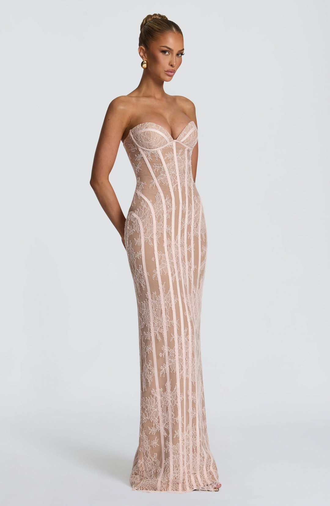 Driekwartsaanzicht van de roze kanten maxi-jurk met lange korsetbanen, model met een hand achter de rug. 16