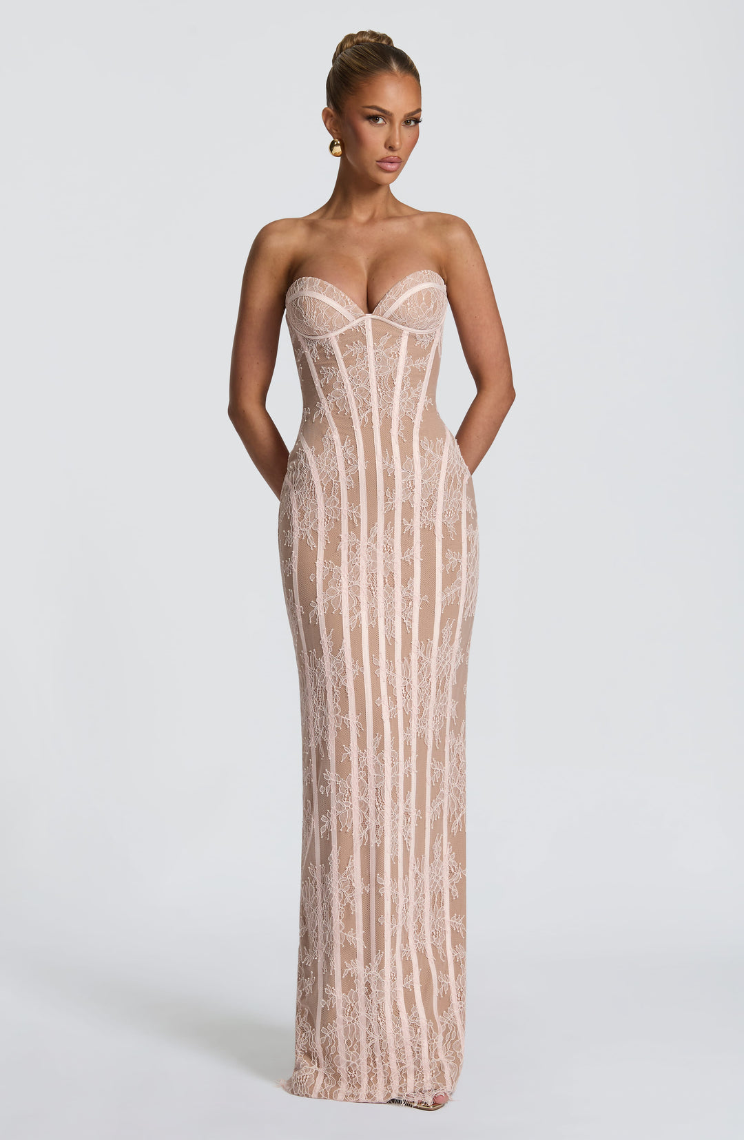 Vooraanzicht van de strapless maxi-jurk van zachtroze kant met korsetbanen, model met opgestoken haar en gouden oorbellen. 13