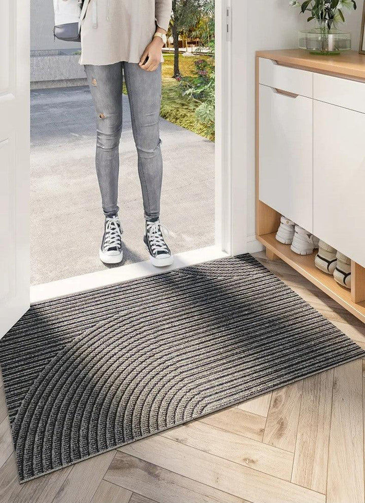 Grijze deurmat met gebogen groeven voor de open voordeur, terwijl iemand in spijkerbroek en sneakers buiten op de drempel staat. 5