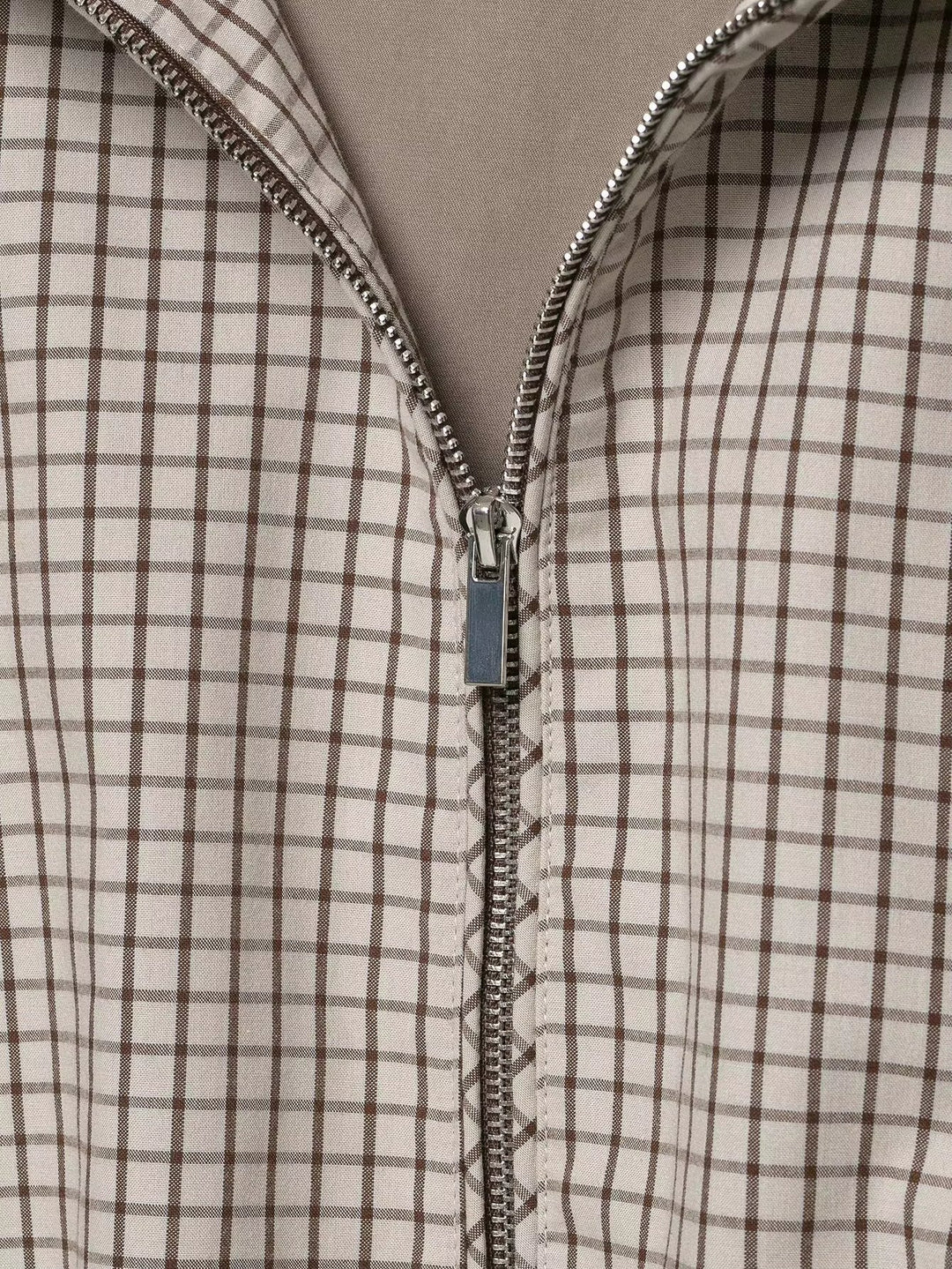 Detail van de metalen rits met ritstrekker, half open, op de geruite stof met beige voering eronder. 6