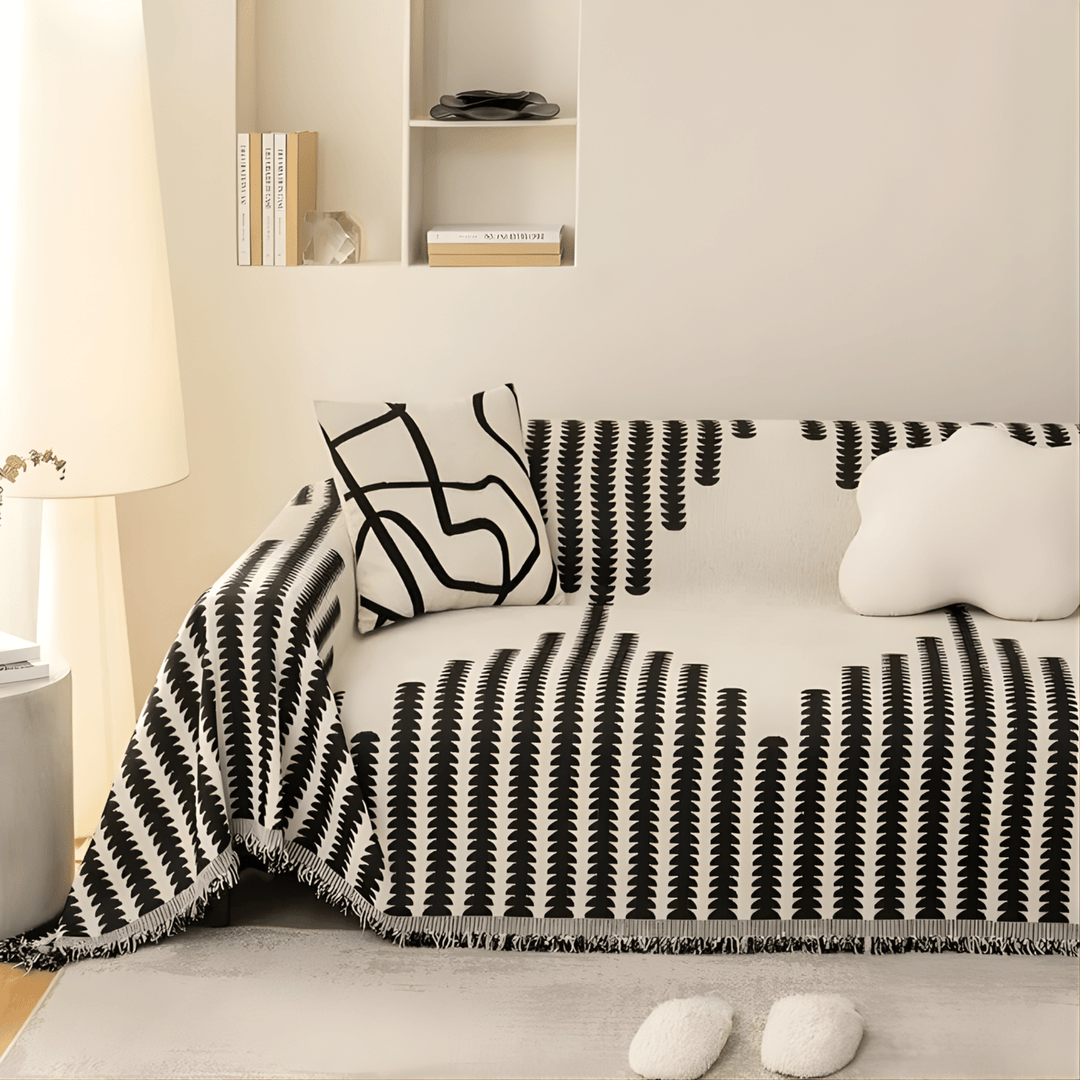 Zwart-wit bankhoes met franjerand over een bank in een lichte woonkamer, met een kussen met lijntekening en een wolkvormig kussen. 3