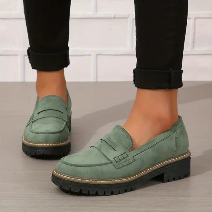 Groene suèdelook loafers met zwarte profielzool, gedragen met zwarte opgerolde broek tegen een witte bakstenen muur. 1