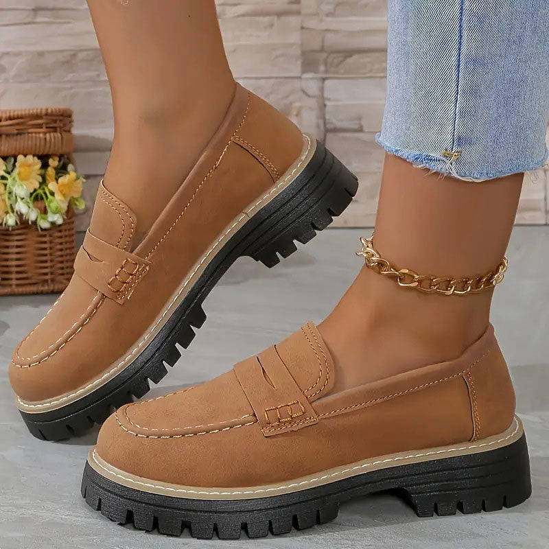Bruine loafers met oranje stiksels en zwarte plateauzool, gedragen met gouden enkelbandje, schuin opgetild getoond. 17