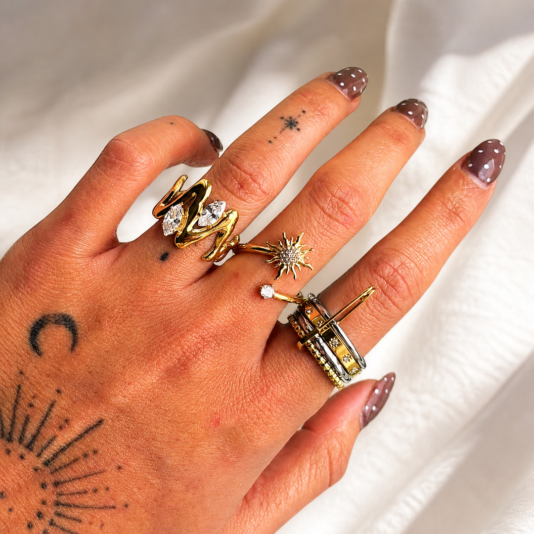 Hand met de goudkleurige golvende ring met twee heldere stenen, gedragen samen met andere ringen, op een witte stoffen ondergrond. 3
