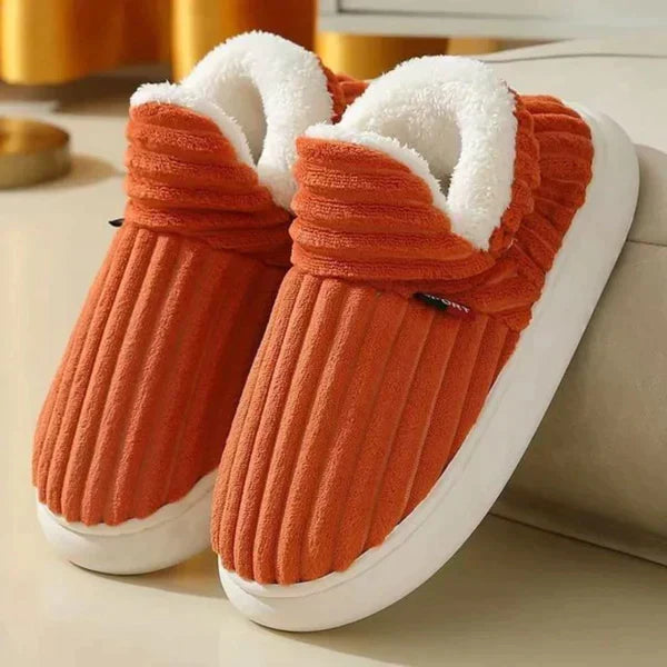 Paar oranje geribbelde pluchen pantoffels met witte fleece voering en dikke witte zool, schuin geplaatst op een lichte vloer. 6