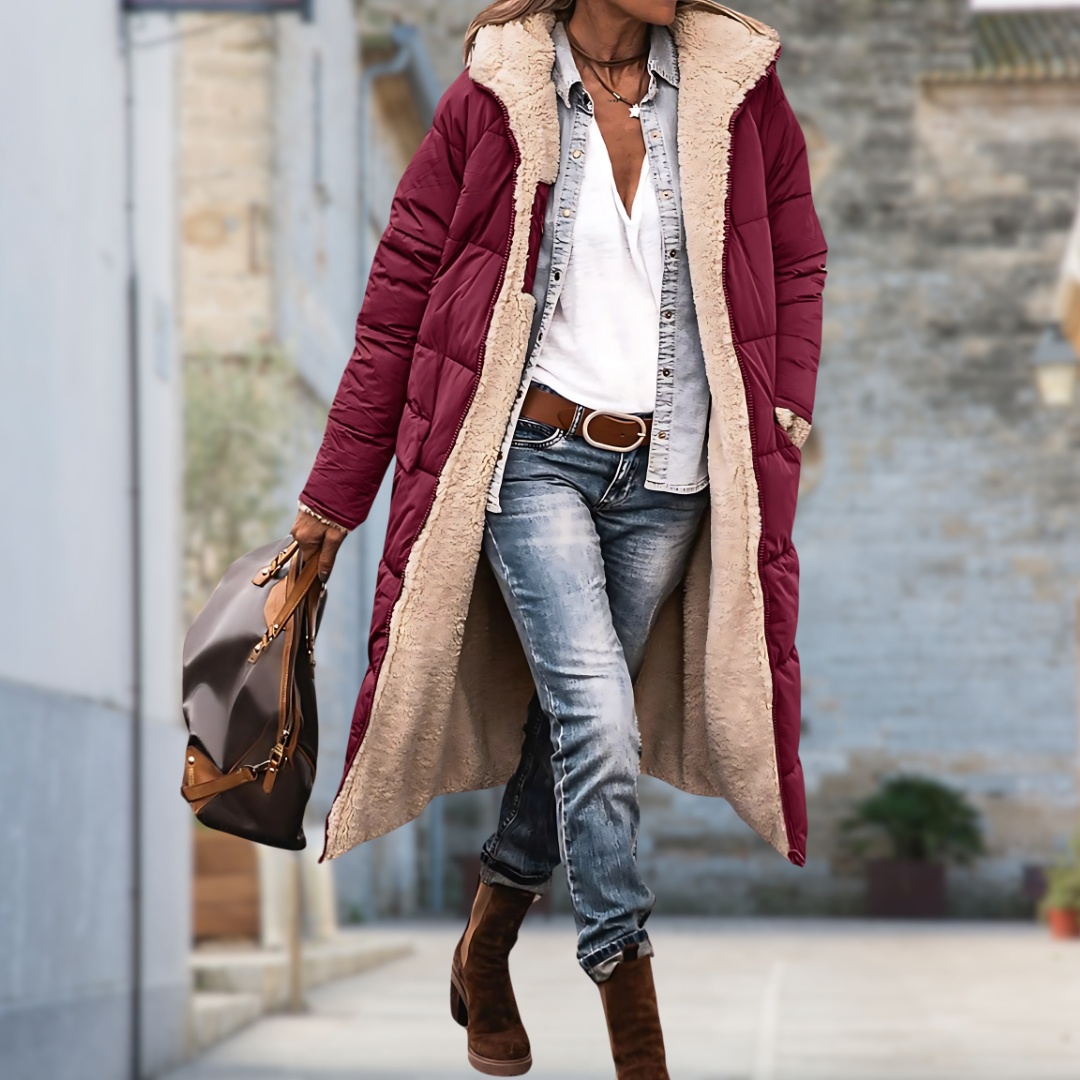 Model loopt door een straat in de lange gewatteerde jas in bordeauxrood, open gedragen met beige fleecevoering aan de binnenkant. 6