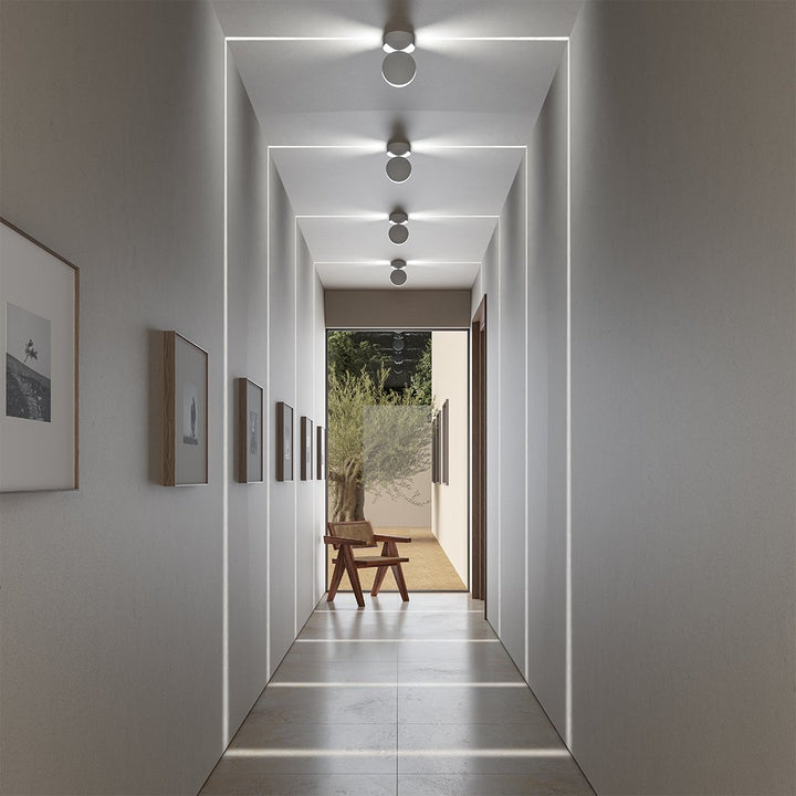 Rij witte plafondlampen in een gang met vierkante lichtlijnen op wanden, plafond en vloer, met een houten stoel aan het einde. 1
