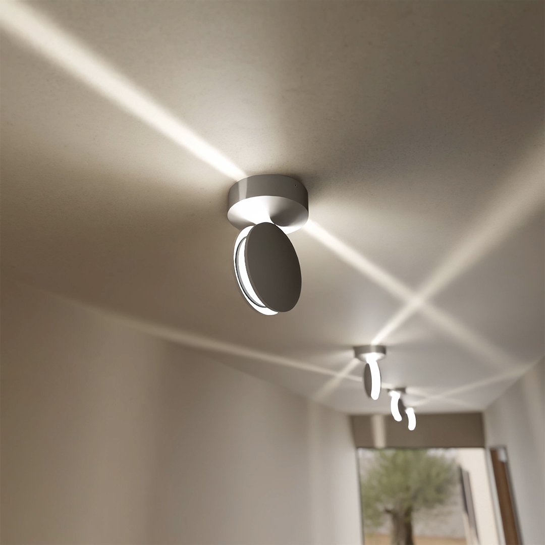 Zilverkleurige plafondlamp met schuin gedraaide kop, die kruisende lichtstralen over het plafond van een gang verspreidt. 4