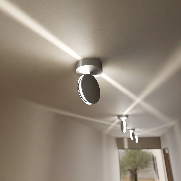 Zilverkleurige plafondlamp met schuin gedraaide kop, die kruisende lichtstralen over het plafond van een gang verspreidt. 4