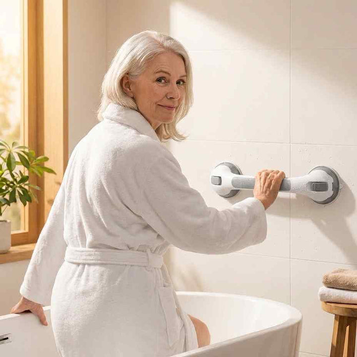 Oudere vrouw in witte badjas stapt een bad in en houdt zich vast aan de wit met grijze beugel op de tegelwand. 1