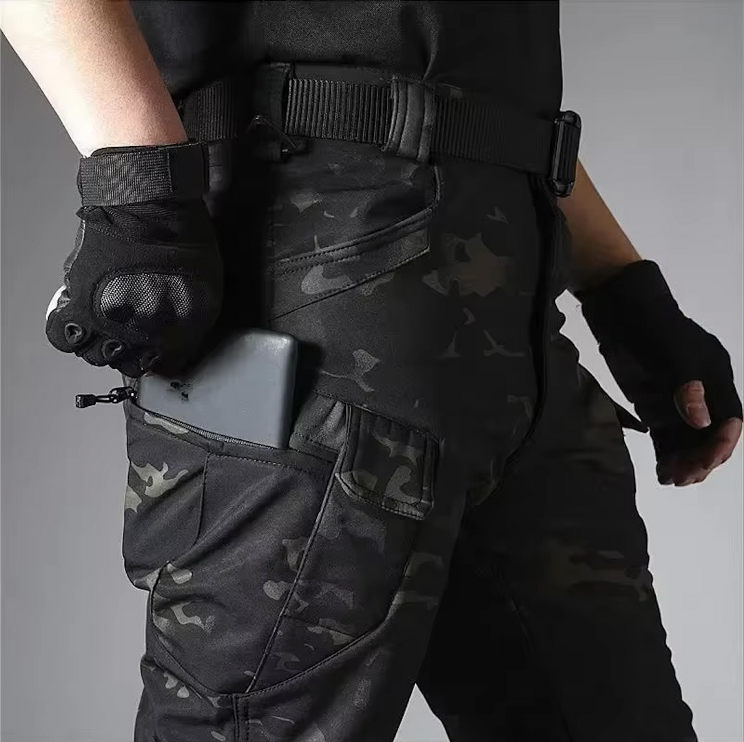 Detail van de broek in donkere camouflageprint: een telefoon in de ritszak van het been, met riem en handschoenen. 19