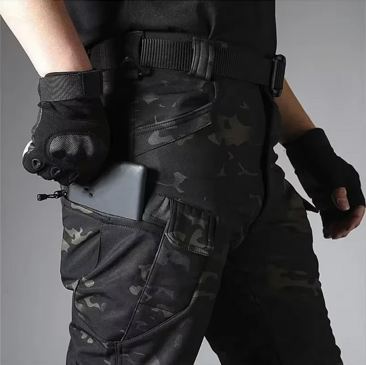 Detail van de broek in donkere camouflageprint: een telefoon in de ritszak van het been, met riem en handschoenen. 19